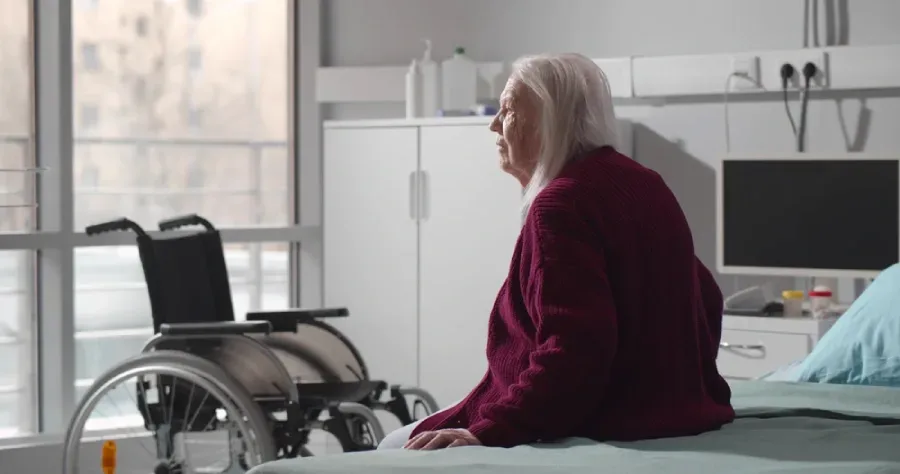 A patient sits on the edge of a hospital bed with a wheelchair in the distance, posing a potential fall risk.
